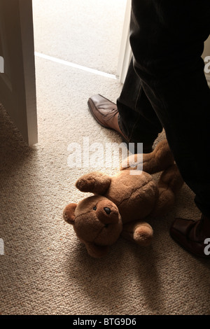 L'uomo (piedi e gambe solo) indossa le scarpe marrone a scavalcare un orsetto sdraiato sul pavimento da una porta aperta. Foto Stock