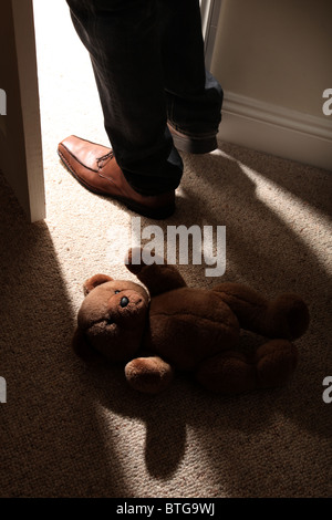 L'uomo (piedi e gambe solo) indossa le scarpe marrone a scavalcare un orsetto sdraiato sul pavimento da una porta aperta. Foto Stock