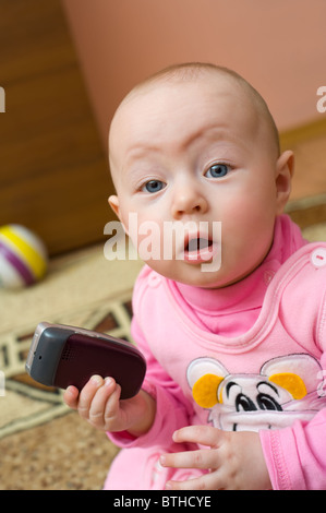 Poco sorpreso europea otto mesi ragazza detiene il telefono cellulare e guarda nella telecamera Foto Stock