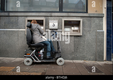 Una donna in una sedia a rotelle motorizzata scooter utilizzando un bancomat, REGNO UNITO Foto Stock