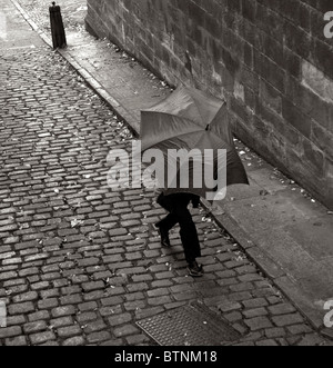 Uomo non identificato che si precipita lungo il cortile acciottolato sotto un ombrello aperto ma in crollo, Lancaster, Regno Unito Foto Stock