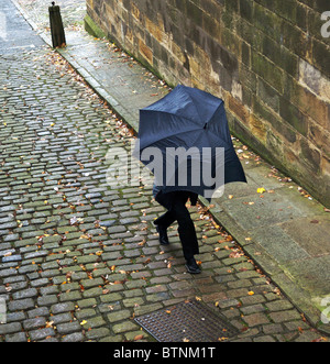 Uomo non identificato che si precipita lungo il cortile acciottolato sotto un ombrello aperto ma in crollo, Lancaster, Regno Unito Foto Stock