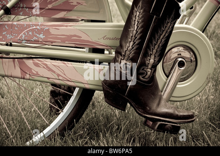 Immagine creativa di una ragazza su una decorativa ladies' bicicletta indossare stivali da cowboy che rappresentano il moderno, nuovo trasporto e i vecchi valori di stile Foto Stock