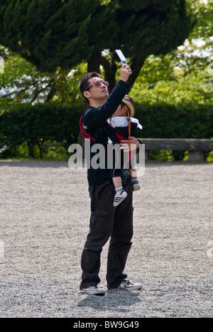 Un padre giapponese tiene il suo bambino in un cablaggio del bambino e prende una foto con il suo cellulare fotocamera Foto Stock