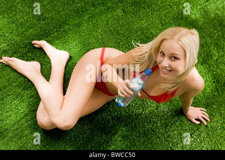 Foto di bella donna bionda seduta sul prato verde con una bottiglia di acqua minerale e guardando la fotocamera Foto Stock