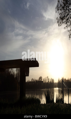 Banco di vuoto che si affaccia su un lago con il sole che tramonta e riflettendo sull'acqua Foto Stock