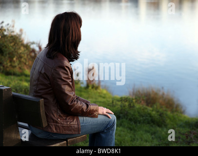 Vista posteriore di una donna nel profondo del pensiero guardando verso un lago, seduta su una panchina nel parco. Foto Stock