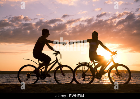 Sagome di felice coppia bracci di estensione per ogni altro mentre in bicicletta sulla riva del mare al tramonto Foto Stock