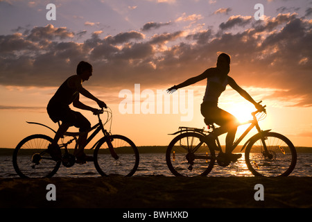 Sagome di coppia felice cavalcare le loro biciclette sulla spiaggia al tramonto Foto Stock