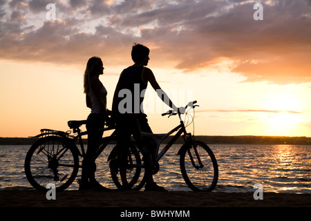 Coppia giovane permanente sulla riva del mare con le loro biciclette e ammirando il tramonto Foto Stock