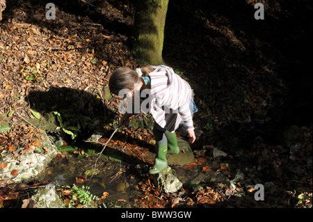 Bambina in verde stivali da pioggia riproduce in un flusso con un bastone tra le foglie di autunno al sole sulla semi-termine holiday Foto Stock