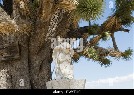 Pregando angelo alato scolpito in pietra decorata con perline e altri elementi, situato a lato strada, Joshua tree in background. Foto Stock