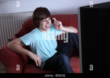 Giovane maschio in possesso di un telefono cellulare sorridente e guardare la tv Foto Stock