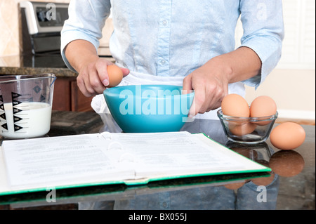 Una massaia crepe un uovo in un recipiente di miscelazione mentre seguendo le istruzioni di un libro di cucina. Foto Stock