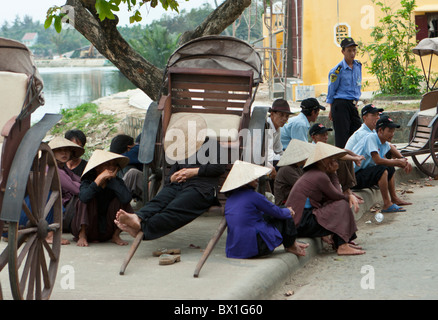 Le donne in cappelli conici, gli uomini nei cappucci, raccolta a riposo su un angolo di strada mentre un rickshaw snoozes driver Foto Stock