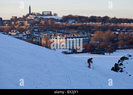 Sciatore in snowy Holyrood Park, Edimburgo, con il Palazzo di Holyrood e Calton Hill al tramonto. Foto Stock