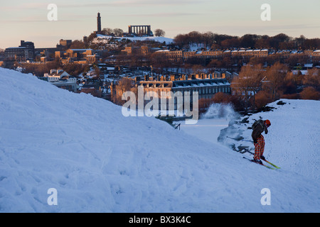 Sciatore in snowy Holyrood Park, Edimburgo, con il Palazzo di Holyrood e Calton Hill al tramonto. Foto Stock