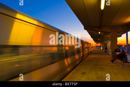 Treno LIRR precipitando in Merrick Stazione ferroviaria al tramonto, donna seduta sul banco della piattaforma sopraelevata, la ferrovia di Long Island, NY Foto Stock