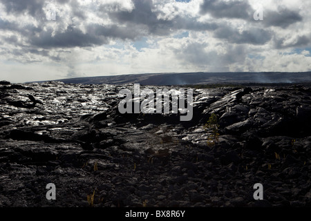 Lava nera formazione sulla Big Island delle Hawaii con cielo blu e nuvole sopra e fumo in background dal flusso di lava Foto Stock