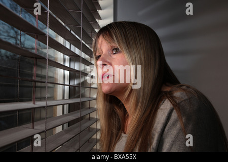 Donna che guarda fuori da una finestra attraverso un cieco Foto Stock