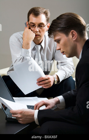 Ritratto di due imprenditore concentrato seduti sulle sedie azienda documenti nelle loro mani e lavorando sul posto per laptop Foto Stock
