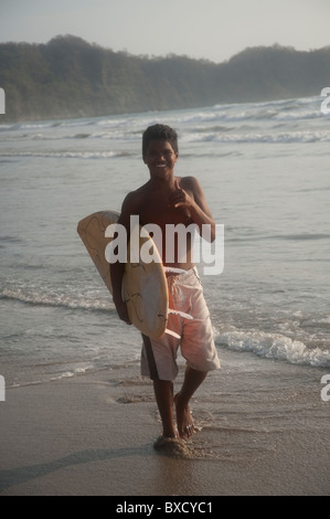 Ragazzo adolescente in un costume da bagno a piedi lungo la spiaggia che porta una tavola da surf Foto Stock