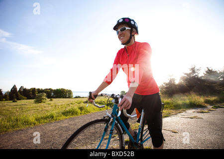 Una giovane donna sorrisi come essa si erge accanto alla sua bicicletta come il sole tramonta sulla sua spalla. Foto Stock