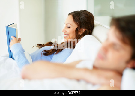 Donna libro di lettura nel letto accanto a dormire marito Foto Stock