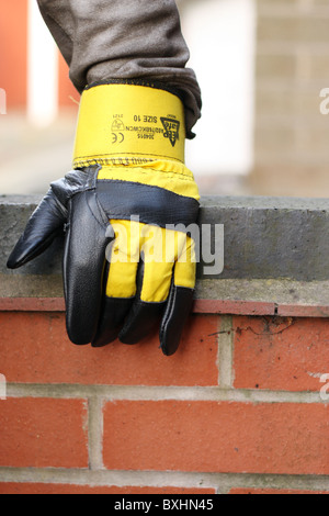 Una mano appoggiata su di un muro di mattoni, indossando un giallo e nero superiore protettivo guanti Rigger. Foto Stock