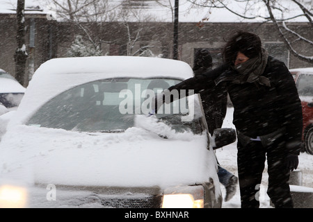 Giovane donna di pulizia vetri auto da neve nella stagione invernale durante la nevicata. Foto Stock