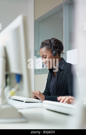 Femmina americano africano rappresentante del servizio clienti a parlare al telefono e digitare su pc. Forma verticale, vista laterale Foto Stock
