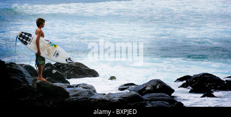 Un giovane surfer guardando il surf break sulle rocce al Parco Hookipa Maui sulle isole Hawaiiano Foto Stock