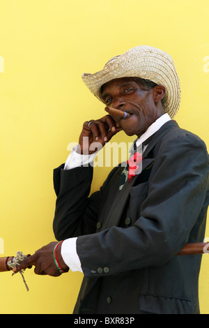 Il vecchio uomo in posa con cappello di paglia, bastone e sigari, contro un giallo brillante parete. Foto Stock