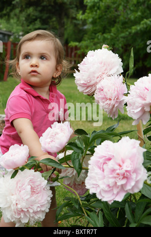 Verticale di un simpatico baby girl scoprendo grandi peonie rosa pallido bloom, che guarda lontano con sentimento misto di espressione. Foto Stock