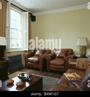 Pelle marrone con poltrone e divani in crema townhouse salotto bianco con persiane in stile piantagione sulla finestra Foto Stock