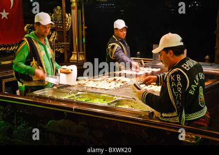 Balik Ekmek Fish Sandwich Vendor Istanbul Turchia // ISTANBUL, Turchia — Un venditore di pesce fresco grigliato su un tradizionale balıkçı teknesi (peschereccio) ormeggiato nelle acque del Corno d'Oro vicino a Eminönü e al Ponte di Galata. Queste cucine galleggianti servono balık ekmek, l'iconico panino di pesce di Istanbul realizzato con sgombro alla griglia servito con pane bianco con cipolle e lattuga. Le barche decorate con decorazioni, spesso caratterizzate da motivi in stile ottomano con finiture dorate e finiture a mezzaluna, sono ormeggiate permanentemente vicino ai terminal dei traghetti di Eminönü. Questa secolare tradizione di venditori di pesce sul lungomare continua ad essere Foto Stock