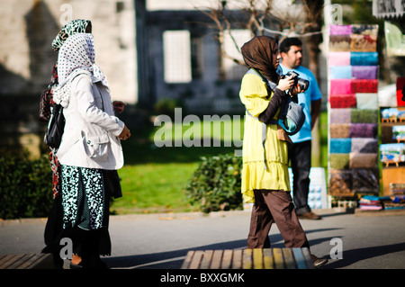 Turisti del Medio Oriente Istanbul Turchia // ISTANBUL, Turchia — turisti del Medio Oriente sono visti esplorare la città di Istanbul, Turchia. Una donna, che indossa un vestito giallo e un hijab, tiene in mano una macchina fotografica per catturare le attrazioni. Altre donne, anche con il velo, camminano nelle vicinanze, mentre un uomo si trova sullo sfondo vicino a un colorato bancone del mercato. Foto Stock