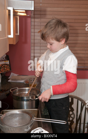 Quattro-anno-vecchio ragazzo con un braccio rotto la cottura fare torte in piedi su una sedia accanto ai fornelli e forno Foto Stock