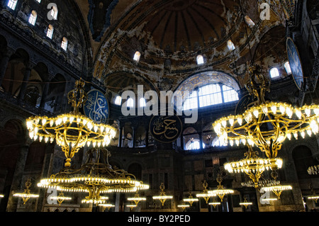 ISTANBUL, Turchia: gli ornati avize (lampadari turchi) sono appesi al soffitto a cupola della Basilica di Santa Sofia, illuminando il vasto spazio interno. Questi apparecchi di illuminazione in bronzo di epoca ottomana sono stati progettati per fornire illuminazione per i servizi di preghiera. L'imponente cupola si innalza a 180 metri dal pavimento, con l'avize sospeso a varie altezze per creare strati di luce. Foto Stock