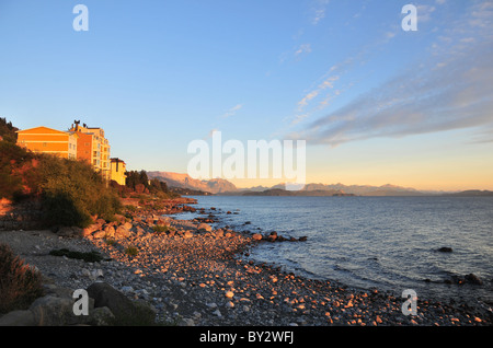 La mattina presto sole che splende su alberghi, spiaggia ghiaia, acqua di lago e sullo sfondo i picchi andini, Lago Nahuel Huapi, Bariloche, Argentina Foto Stock