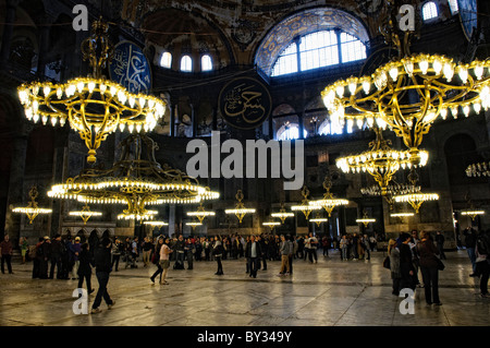 ISTANBUL, Turchia: gli ornati avize (lampadari turchi) sono appesi al soffitto a cupola della Basilica di Santa Sofia, illuminando il vasto spazio interno. Questi apparecchi di illuminazione in bronzo di epoca ottomana sono stati progettati per fornire illuminazione per i servizi di preghiera. L'imponente cupola si innalza a 180 metri dal pavimento, con l'avize sospeso a varie altezze per creare strati di luce. Foto Stock