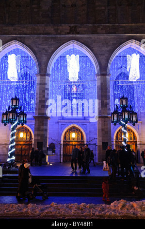 Basilique Notre-Dame de Montreal luci di Natale di notte con persone | MONTREAL, Quebec, Canada - angeli illuminati e cascate di luci blu riempiono i tre archi in stile neogotico della basilica Notre-Dame de Montréal in una serata invernale mentre i visitatori si riuniscono sulla piazza innevata. La facciata ad arco a punta, completata nel 1829 su progetto dell'architetto irlandese James o'Donnell, introdusse l'architettura neogotica in Canada decenni prima che lo stile si diffondesse in tutto il Nord America. Foto Stock