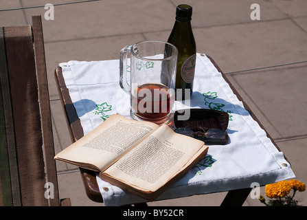 Aprire il libro e gli occhiali da lettura sul piccolo tavolo in legno con una bottiglia di Guinness accanto a una vecchia sedia a sdraio Foto Stock