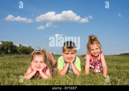 Due gemelle, 3 anni, e il loro fratello, 7 anni, all'aperto, giacente insieme sul campo di erba sotto il cielo blu con nuvole Foto Stock