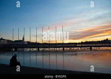 Un ragazzo si siede dal laghetto congelato in Reykjavík, Islanda, al tramonto. Foto Stock