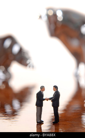 Due manager figurine stringono le mani, con bull e bear fuori fuoco dietro di loro, immagine simbolica per stock exchange business Foto Stock