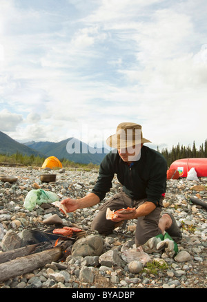 L'uomo la cottura su un fuoco di campo, barbeque, grigliare hamburger, ghiaia bar, tenda dietro, camping, Wind River, Yukon Territory, Canada Foto Stock