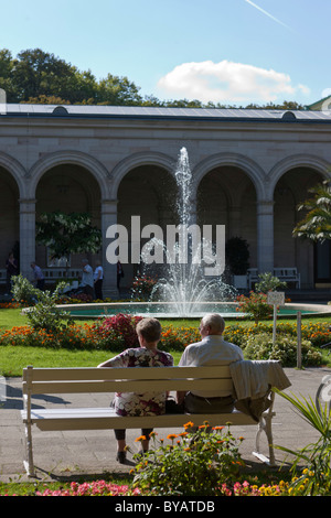 I pensionati su una panchina nel parco di fronte Kurhaus spa hotel con l'edificio Regentenbau e sale arcade, Kurgarten, spa garden Foto Stock