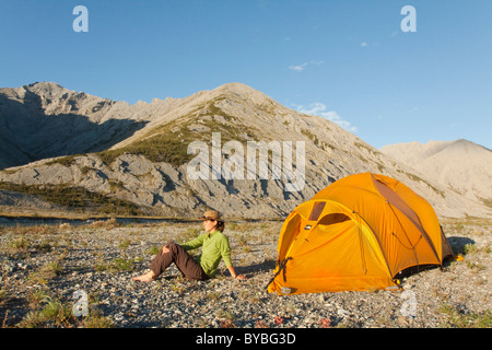 Giovane donna seduta, rilassante, gustando la luce della sera, expedition tenda, tundra artica, camping, Mackenzie montagne dietro Foto Stock