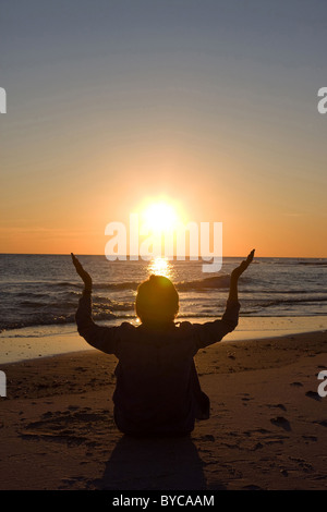 Donna matura si siede sulla spiaggia con le mani alzate come il sole tramonta sull'acqua. Camera per copiare in alto. Foto Stock